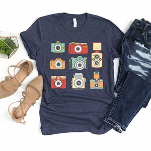 May include: A navy blue t-shirt with a graphic of nine vintage cameras in various colors and styles. The cameras are arranged in a grid pattern on the shirt.