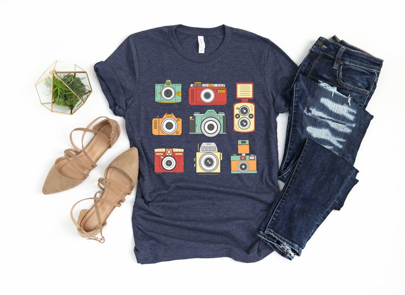 May include: A navy blue t-shirt with a graphic of nine vintage cameras in various colors and styles. The cameras are arranged in a grid pattern on the shirt.