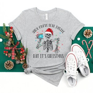May include: A gray t-shirt with a skeleton holding a mug of hot chocolate. The text on the shirt reads "When you're dead inside but it's Christmas".