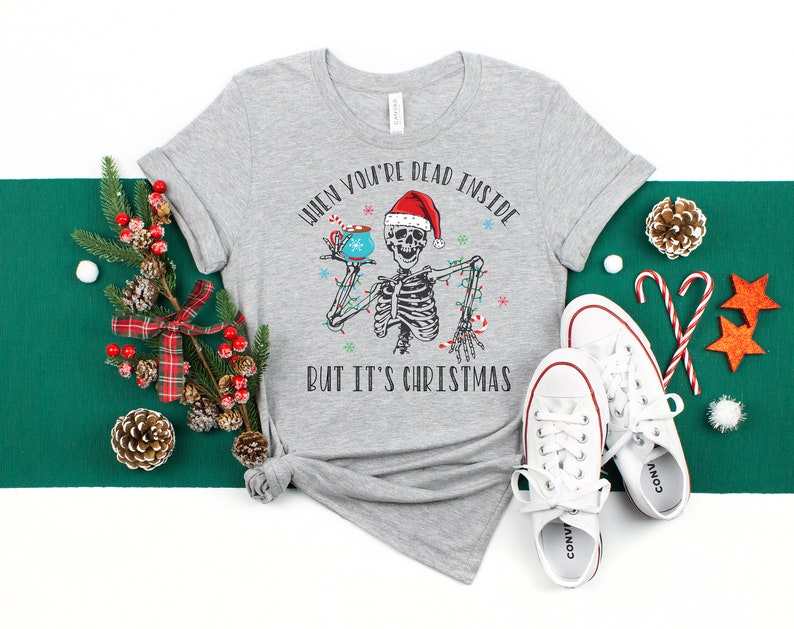 May include: A gray t-shirt with a skeleton holding a mug of hot chocolate. The text on the shirt reads "When you're dead inside but it's Christmas".