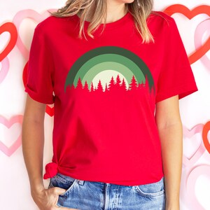 May include: A red t-shirt with a green and white forest silhouette design. The design features a stylized rainbow arch over the trees.