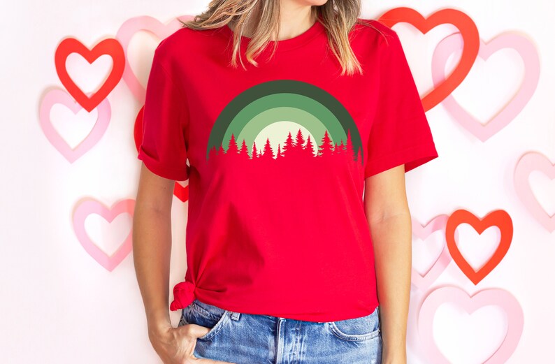 May include: A red t-shirt with a green and white forest silhouette design. The design features a stylized rainbow arch over the trees.
