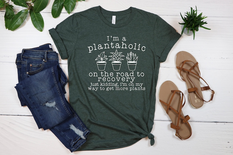 May include: A green t-shirt with the text "I'm a plantaholic on the road to recovery just kidding, I'm on my way to get more plants". The text is in white and there are three small drawings of potted plants.