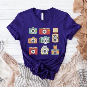 May include: Purple t-shirt with a vintage camera design. The design features eight different cameras in various colors and styles.