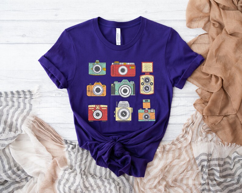May include: Purple t-shirt with a vintage camera design. The design features eight different cameras in various colors and styles.