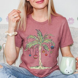 May include: A woman wearing a mauve t-shirt with a palm tree design and colorful ornaments. The palm tree is decorated with ornaments that resemble Christmas ornaments. The woman is holding a light green mug with a white rabbit design.