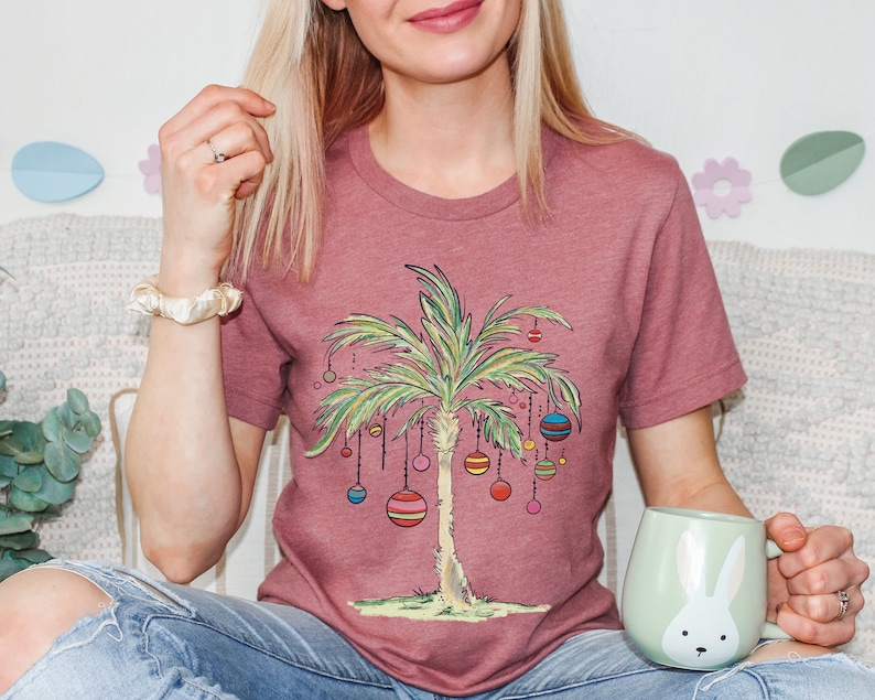 May include: A woman wearing a mauve t-shirt with a palm tree design and colorful ornaments. The palm tree is decorated with ornaments that resemble Christmas ornaments. The woman is holding a light green mug with a white rabbit design.