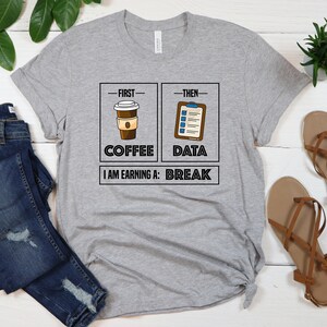 May include: Gray t-shirt with a graphic of a coffee cup and a checklist. The text reads "First - Coffee, Then - Data, I am earning a: Break".