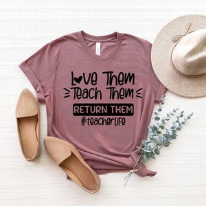 May include: A mauve t-shirt with the text "Love Them Teach Them Return Them #teacherlife" printed in black. The shirt has a heart symbol above the word "Love".