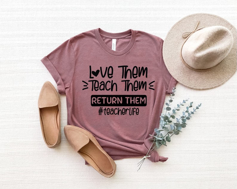 May include: A mauve t-shirt with the text "Love Them Teach Them Return Them #teacherlife" printed in black. The shirt has a heart symbol above the word "Love".