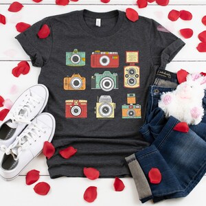 May include: A dark gray t-shirt with a vintage camera design. The shirt features a variety of cameras in different colors and styles. The shirt is laid out on a white wooden surface with white sneakers, blue jeans, and a white stuffed bunny.