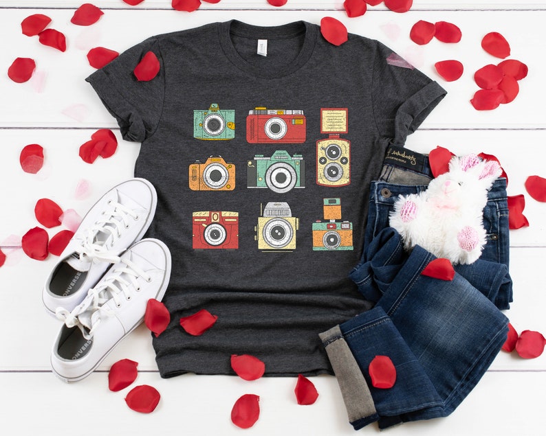 May include: A dark gray t-shirt with a vintage camera design. The shirt features a variety of cameras in different colors and styles. The shirt is laid out on a white wooden surface with white sneakers, blue jeans, and a white stuffed bunny.
