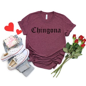 Op de afbeelding: Een maroon t-shirt met het woord "Chingona" gedrukt in zwarte gotische letters.