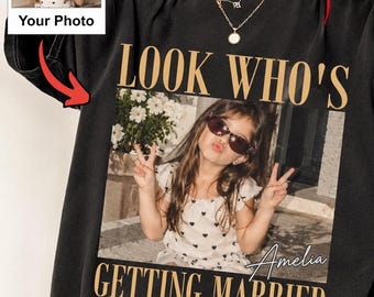 May include: A black crewneck t-shirt featuring a custom photo print of a child wearing sunglasses and making a peace sign. The text reads LOOK WHO'S GETTING MARRIED with the name Amelia in script. A small inset shows the original photo source.