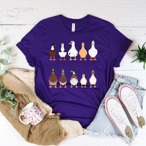 May include: A purple t-shirt with a graphic of a line of ducks in different colors and breeds. The ducks are facing forward and have their heads up.