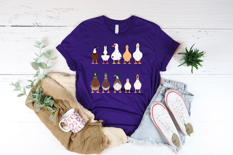 May include: A purple t-shirt with a graphic of a line of ducks in different colors and breeds. The ducks are facing forward and have their heads up.