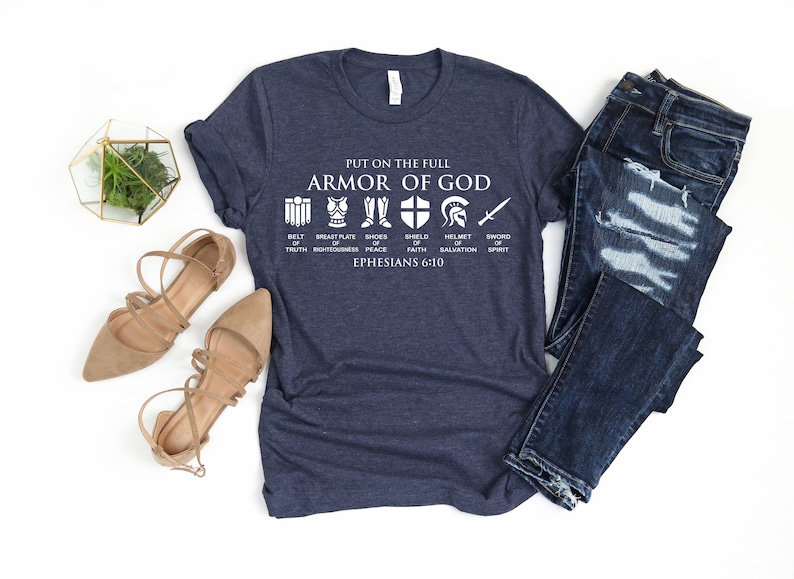 May include: A navy blue t-shirt with a white graphic of the armor of God. The graphic includes icons representing the belt of truth, breastplate of righteousness, shoes of peace, shield of faith, helmet of salvation, and sword of the spirit. The text "PUT ON THE FULL ARMOR OF GOD" and "EPHESIANS 6:10" are also included in the graphic.