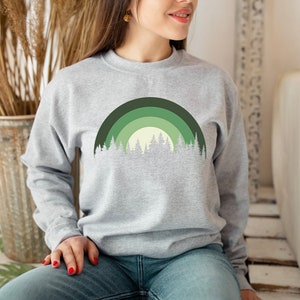 May include: A gray sweatshirt with a green and white forest design. The design features a stylized rainbow with trees in front of it.