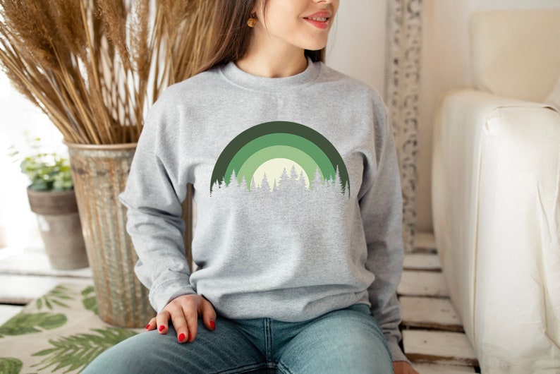 May include: A gray sweatshirt with a green and white forest design. The design features a stylized rainbow with trees in front of it.