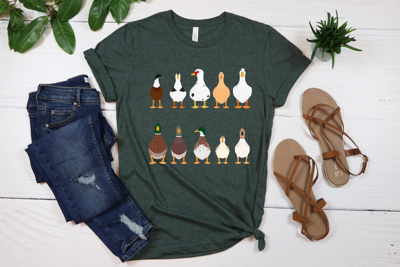 May include: A dark green t-shirt with a graphic of nine ducks in a row. The ducks are all facing away from the viewer and have different colored feathers. The t-shirt is on a white wooden surface.