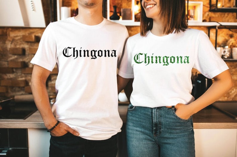 Op de afbeelding: Twee witte T-shirts met het woord "Chingona" erop gedrukt in zwarte en groene letters. De shirts worden gedragen door twee mensen.