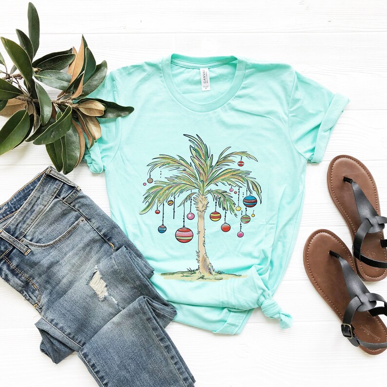 May include: A turquoise t-shirt with a palm tree decorated with colorful ornaments. The palm tree is a Christmas tree design. The t-shirt is laid out on a white surface with a pair of brown sandals and blue jeans.