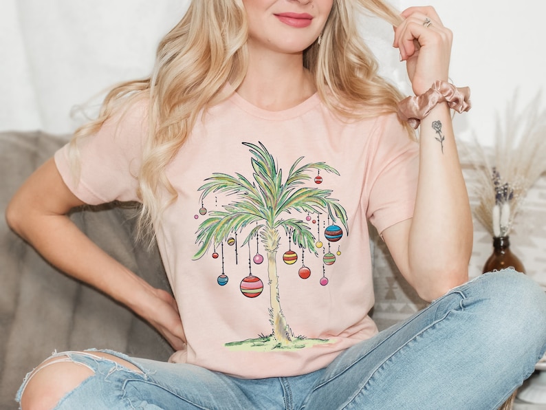 May include: A pink t-shirt with a graphic of a palm tree decorated with colorful ornaments. The palm tree is a whimsical take on a traditional Christmas tree.