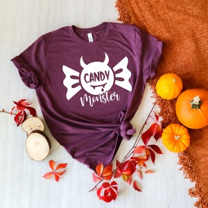 May include: Purple t-shirt with a white graphic of a candy with devil horns and the text "Candy Monster". The shirt is laid out on a white wooden surface with fall leaves and pumpkins.