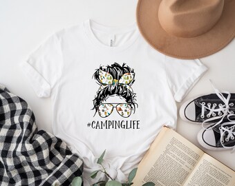 May include: White t-shirt with a black and white illustration of a woman with a messy bun. The bun is decorated with camping-themed patterns. The woman is wearing sunglasses with the same camping-themed patterns. The text "#CAMPINGLIFE" is printed below the illustration.