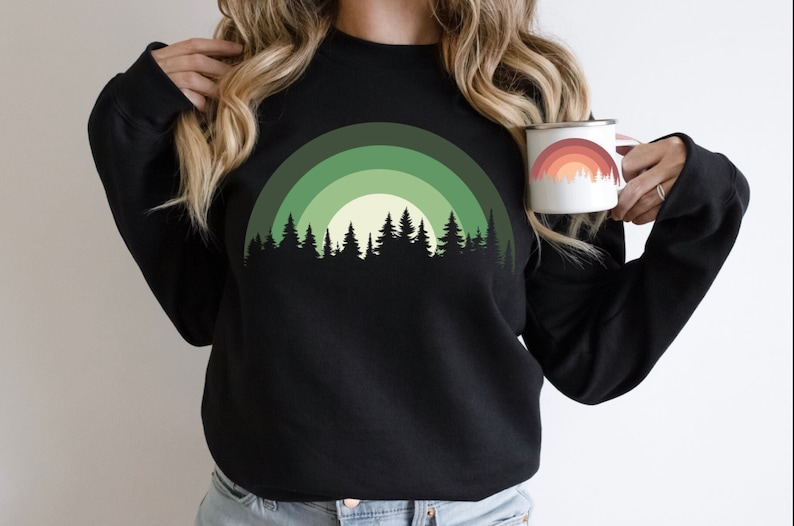 May include: Black sweatshirt with a green and white forest silhouette design. The design features a half-circle of trees with a sun behind them.