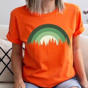 May include: Orange short-sleeved t-shirt with a green and white forest silhouette design. The design features a crescent moon shape with a row of trees at the bottom.