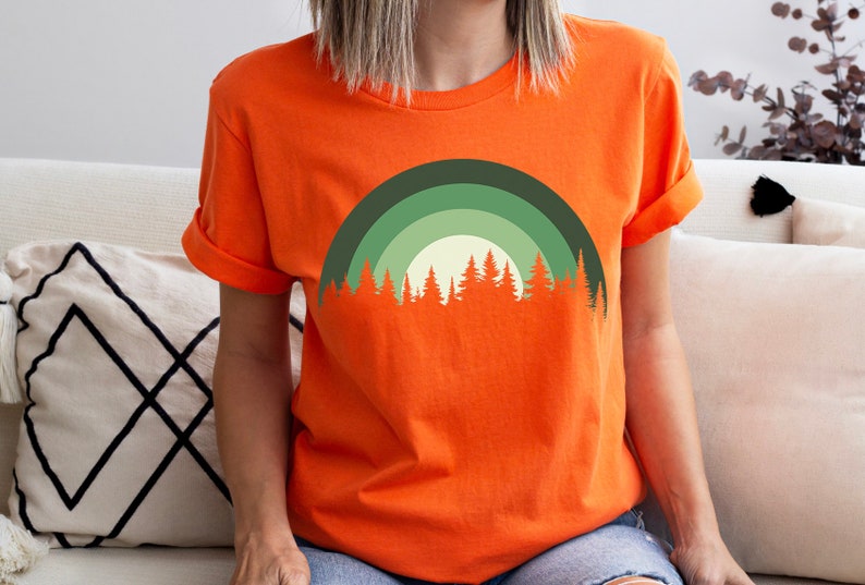 May include: Orange short-sleeved t-shirt with a green and white forest silhouette design. The design features a crescent moon shape with a row of trees at the bottom.