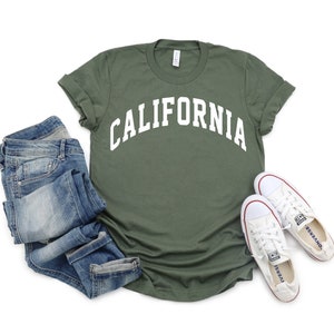 Peut inclure: Un t-shirt vert avec le mot "CALIFORNIA" imprim&eacute; en blanc sur le devant. Le t-shirt est pos&eacute; &agrave; plat sur un fond blanc avec un jean bleu et des baskets blanches.