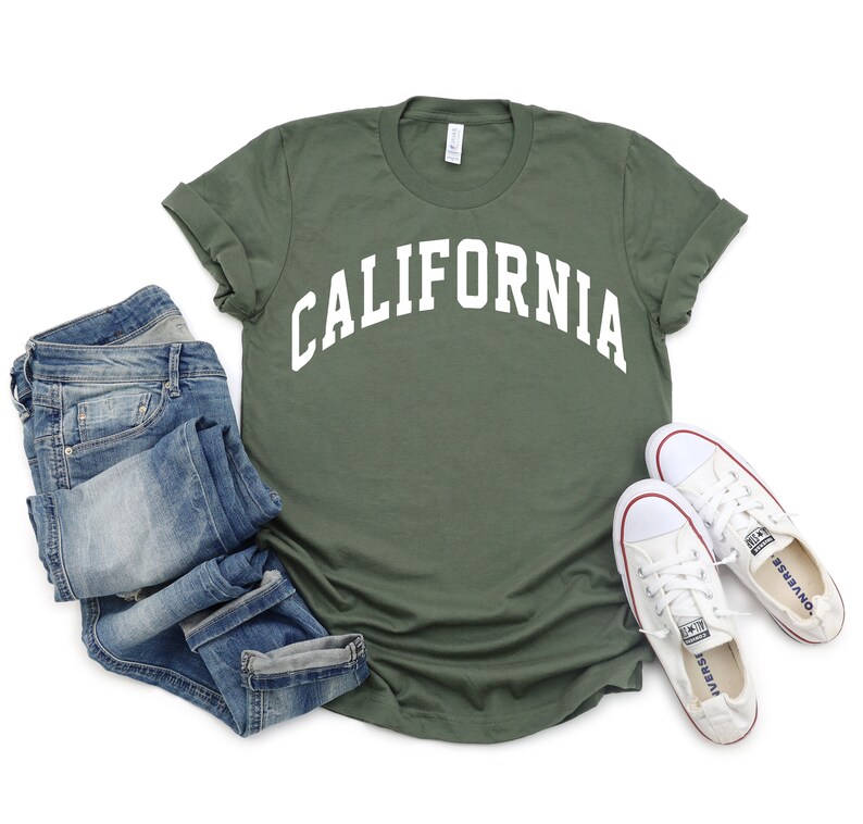 Peut inclure: Un t-shirt vert avec le mot "CALIFORNIA" imprim&eacute; en blanc sur le devant. Le t-shirt est pos&eacute; &agrave; plat sur un fond blanc avec un jean bleu et des baskets blanches.