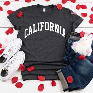 Peut inclure: Un t-shirt gris fonc&eacute; avec le mot "CALIFORNIA" imprim&eacute; en blanc sur le devant. Le t-shirt est pos&eacute; sur une surface blanche avec des p&eacute;tales de roses rouges &eacute;parpill&eacute;s autour. Il y a aussi une paire de baskets blanches, un jean bleu et un lapin en peluche blanc sur la surface.