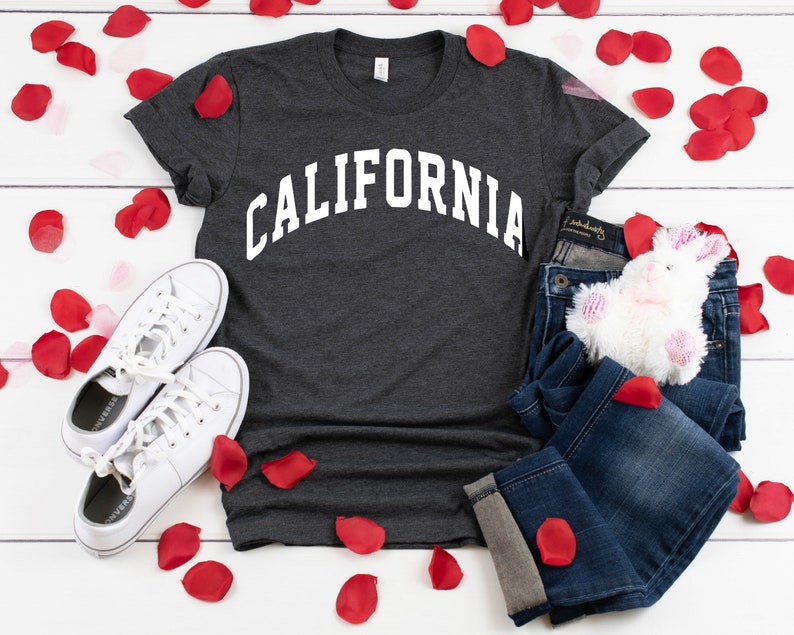 Peut inclure: Un t-shirt gris fonc&eacute; avec le mot "CALIFORNIA" imprim&eacute; en blanc sur le devant. Le t-shirt est pos&eacute; sur une surface blanche avec des p&eacute;tales de roses rouges &eacute;parpill&eacute;s autour. Il y a aussi une paire de baskets blanches, un jean bleu et un lapin en peluche blanc sur la surface.