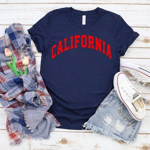 Peut inclure: Un t-shirt bleu marine avec le mot "CALIFORNIA" imprim&eacute; en rouge sur le devant. Le t-shirt est pos&eacute; sur une surface en bois blanc avec une chemise &agrave; carreaux, des fleurs et des baskets blanches.