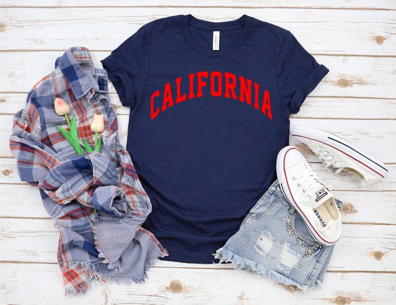 Peut inclure: Un t-shirt bleu marine avec le mot "CALIFORNIA" imprim&eacute; en rouge sur le devant. Le t-shirt est pos&eacute; sur une surface en bois blanc avec une chemise &agrave; carreaux, des fleurs et des baskets blanches.
