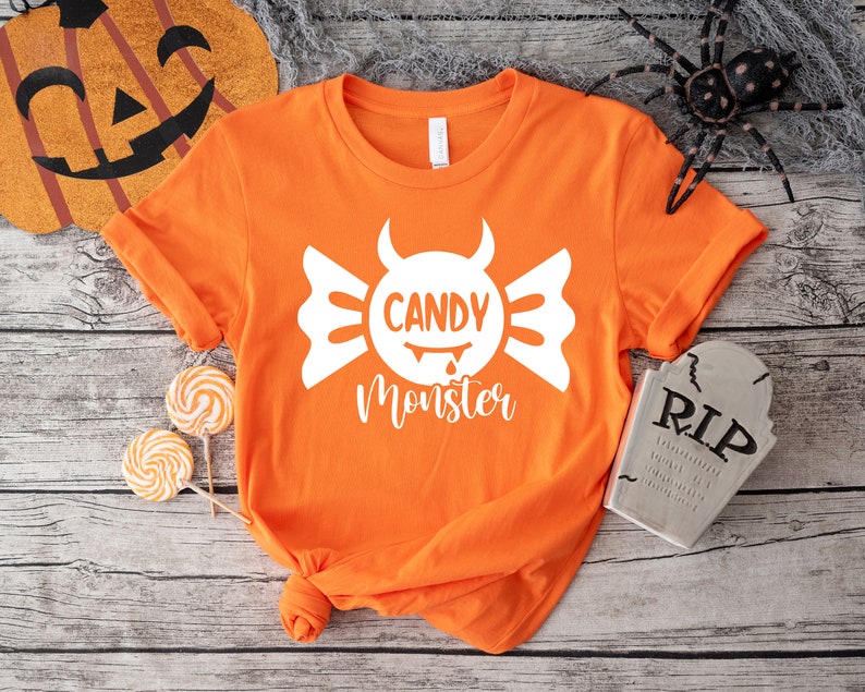 May include: Orange t-shirt with a white candy monster graphic. The graphic features a white candy with a devil's horns and the text "Candy Monster".