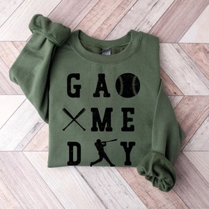 May include: A green sweatshirt with the text "GAME DAY" in black lettering. The text is arranged vertically with a baseball, crossed bats, and a baseball player graphic.