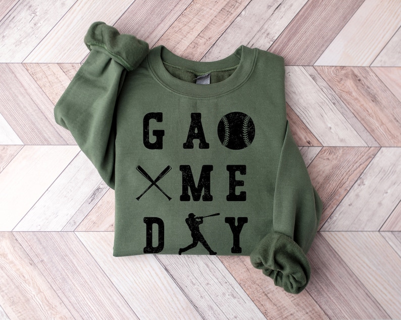 May include: A green sweatshirt with the text "GAME DAY" in black lettering. The text is arranged vertically with a baseball, crossed bats, and a baseball player graphic.