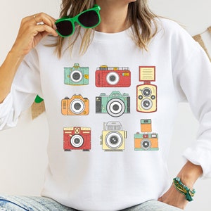 May include: White sweatshirt with a vintage camera design. The cameras are in various colors, including red, green, blue, and yellow.