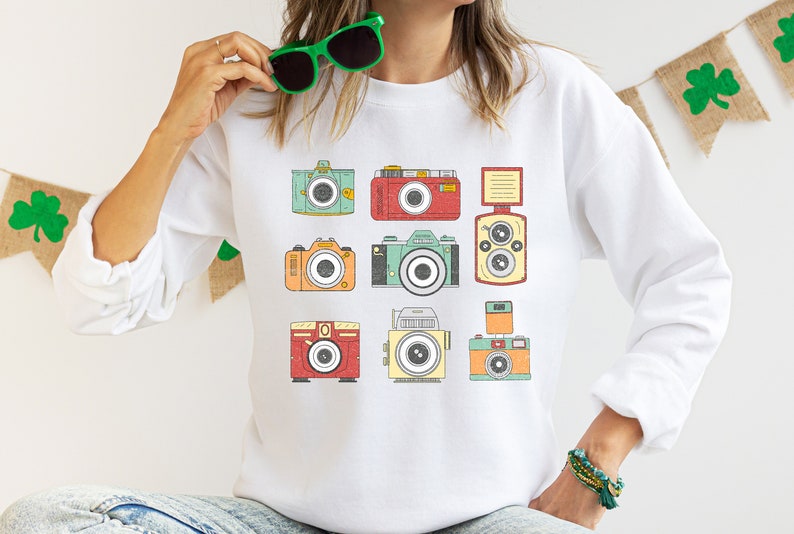 May include: White sweatshirt with a vintage camera design. The cameras are in various colors, including red, green, blue, and yellow.