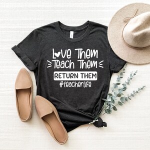 May include: A dark gray t-shirt with the text "Love Them Teach Them Return Them #teacherlife" printed in white. The shirt is laid flat on a white surface with a brown hat and two pairs of brown shoes.