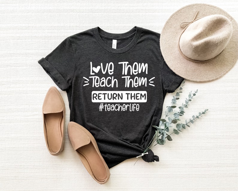 May include: A dark gray t-shirt with the text "Love Them Teach Them Return Them #teacherlife" printed in white. The shirt is laid flat on a white surface with a brown hat and two pairs of brown shoes.