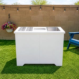 May include: A white rectangular outdoor serving station featuring a flat countertop with six small circular stainless steel inserts, one rectangular stainless steel tray, and one larger square stainless steel basin integrated into the top surface.