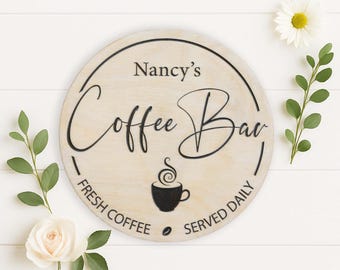 May include: A round wooden sign with the text "Nancy's Coffee Bar" in black script. The sign also features a black coffee cup graphic and the words "Fresh Coffee Served Daily". The sign is surrounded by white flowers and green leaves.
