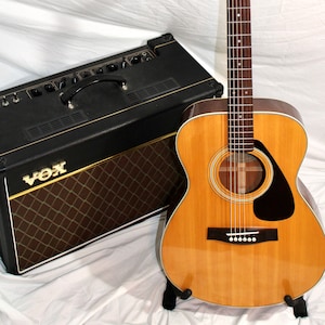 Puede incluir: Una guitarra acústica marrón con una golpeador negro y clavijas cromadas. La guitarra tiene la palabra "YAMAHA" impresa en la pala. La guitarra está sentada en un soporte delante de un amplificador negro con la palabra "VOX" impresa en la parte delantera.