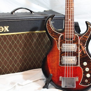 Può includere: Un amplificatore Vox vintage con una griglia a rombi marroni e un logo VOX dorato. Un basso elettrico rosso, nero e marrone con una forma del corpo unica e due pickup è davanti all'amplificatore.