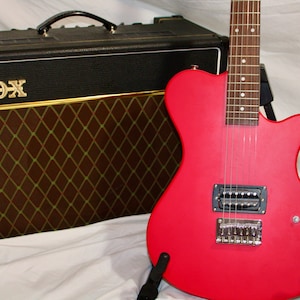 Puede incluir: Una guitarra eléctrica roja con una golpeador negro y un diapasón de madera marrón. La guitarra está en un soporte delante de un amplificador Vox negro.